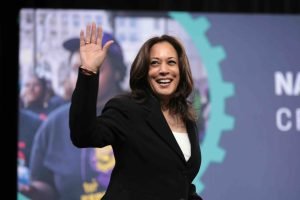 U.S. Senator Kamala Harris speaking with attendees at the 2019 National Forum on Wages and Working People hosted by the Center for the American Progress Action Fund and the SEIU at the Enclave in Las Vegas, Nevada. She is wearing a black suit, white top, and is stood on stage. She's pictured waving to the audience and smiling.