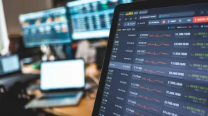 A close-up shot of a crypto exchange screen with various cryptocurrencies listed. The screen is displaying the current prices of Bitcoin, Ethereum, and several other cryptocurrencies. The background is blurred, showing a room with multiple monitors and a laptop.